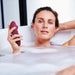 A woman relaxes in a bubble bath, holding a Womanizer Classic 2 Pleasure Air Clitoral Stimulator - Bordeaux in her hand. She has a content expression on her face and leans back with one arm resting on the tub edge. The background is mostly white with soft lighting.