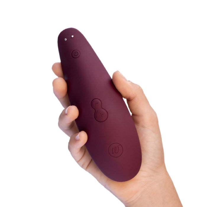 A hand holding a bordeaux, oval-shaped electronic device with multiple buttons. This Womanizer Classic 2 Pleasure Air Clitoral Stimulator by Womanizer features air pulsation technology and has a smooth surface with three control buttons, including a power button and plus/minus buttons for adjustment, designed specifically for clitoral stimulation.