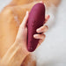 A hand holding the Womanizer Classic 2 Pleasure Air Clitoral Stimulator in Bordeaux, featuring waterproof capabilities and physical control buttons, being used in a bubble bath for ultimate clitoral stimulation.