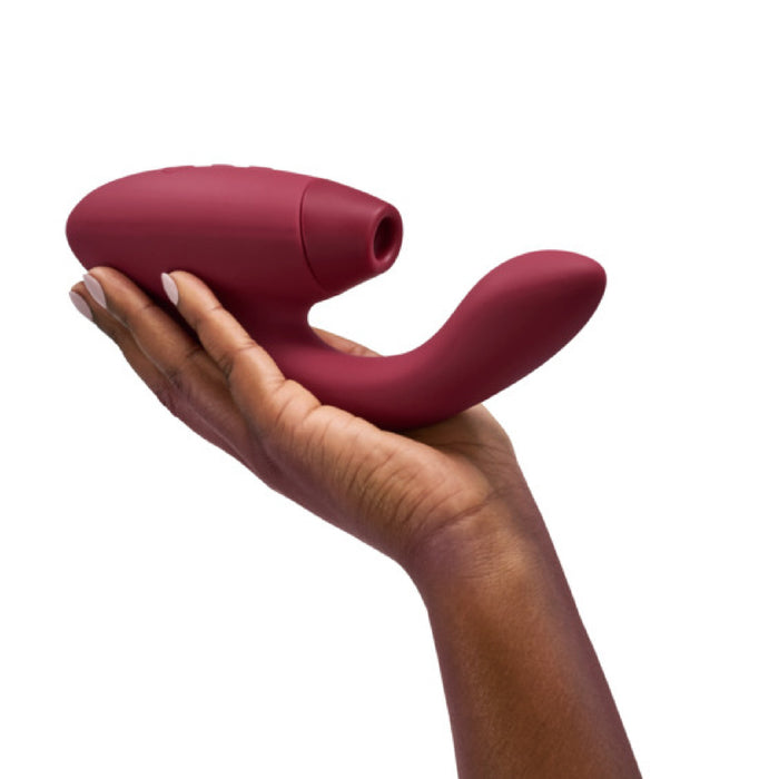 A hand holds the Womanizer Duo 2 Pleasure Air Clitoral & G-Spot Rabbit in Bordeaux, showcasing its curved handle and nozzle-like opening for clitoral air pulsation. The plain white background highlights the sleek and smooth design of this customizable pleasure device by Womanizer.