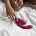 A person with medium brown skin sits on a bed with white sheets, reaching toward a Bordeaux-colored Womanizer Duo 2 Pleasure Air Clitoral & G-Spot Rabbit. Their hand is positioned near the clitoral air pulsation device, and their legs are partially visible in the background.