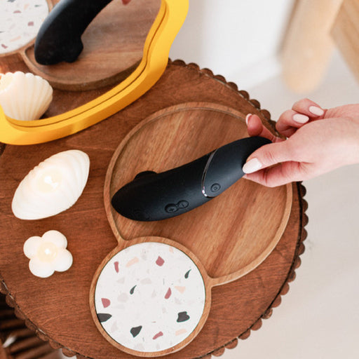 A hand holding a Womanizer Next Pleasure Air Clitoral Vibrator in black above a wooden table. The table features a small yellow mirror, two decorative candles shaped like a shell and a flower, and a round placemat with terrazzo design.