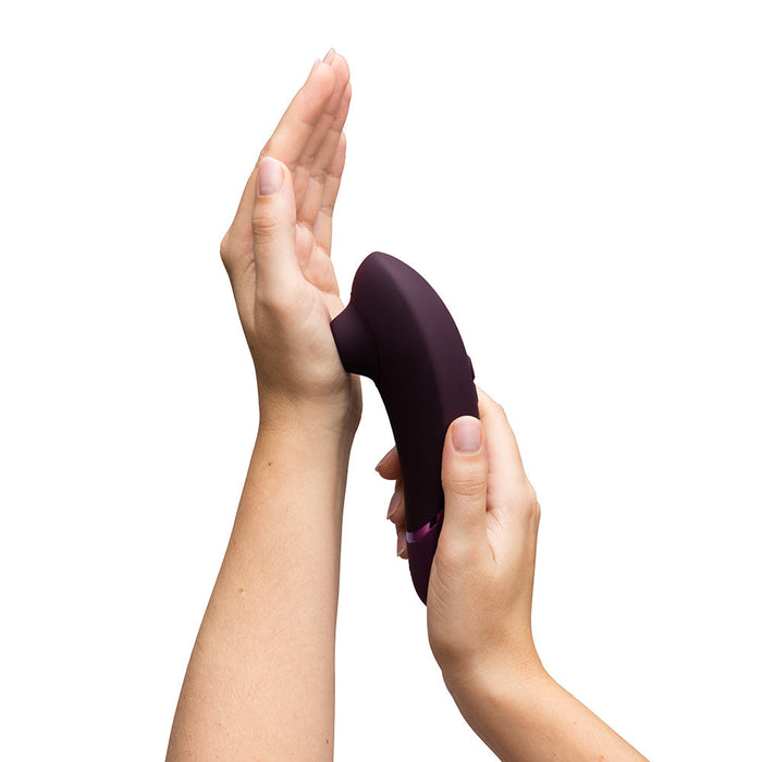 A person holds a plum-colored, ergonomically designed Womanizer Next Pleasure Air Clitoral Vibrator by Womanizer, pressing it against the palm of their other hand with fingers curved around its contoured shape. The innovative Pleasure Air Technology promises unparalleled clitoral stimulation. The background is white.