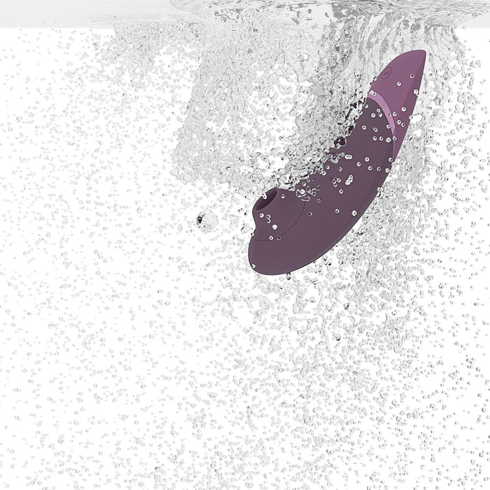 The Womanizer Next Pleasure Air Clitoral Vibrator - Plum, a purple vibrating personal care device, is shown submerged in water with numerous bubbles and splashes surrounding it, demonstrating its waterproof feature. The clear liquid and dynamic motion evoke a sense of the device being used in a wet environment with its innovative Pleasure Air Technology.