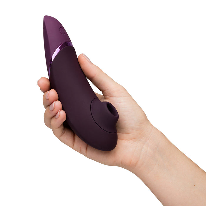 A hand holding a Womanizer Next Pleasure Air Clitoral Vibrator in Plum, exhibiting a sleek, curved design with a circular opening at the tip, suggesting it’s likely a personal massager. The device has a glossy finish with a metallic accent near the base and is equipped with Pleasure Air Technology for superior comfort.