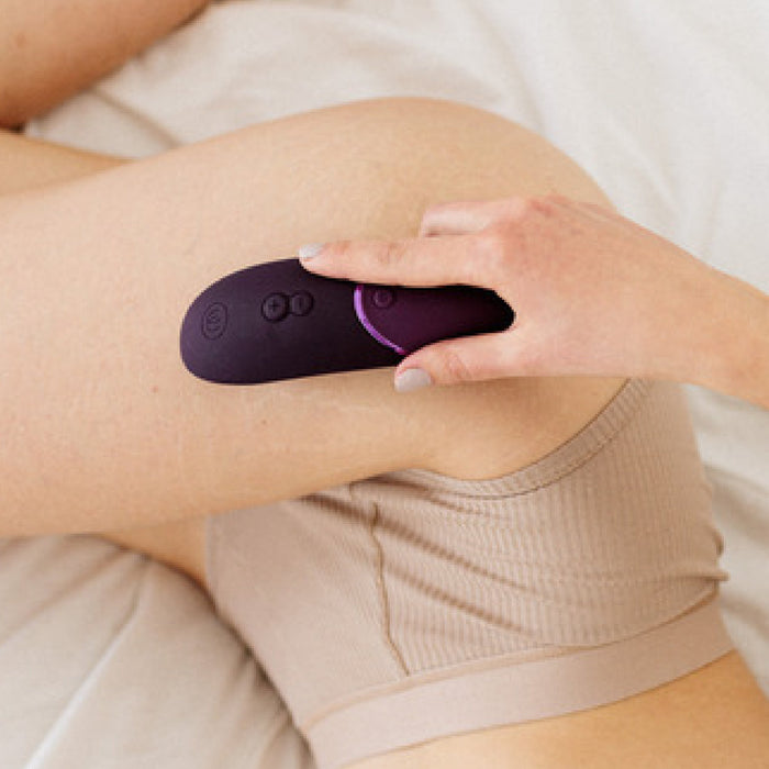 A close-up image of a person wearing beige shorts holding a plum-colored Womanizer Next Pleasure Air Clitoral Vibrator by Womanizer against their thigh. The person is resting on a light-colored surface. The device appears to feature advanced Pleasure Air Technology for enhanced user experience.