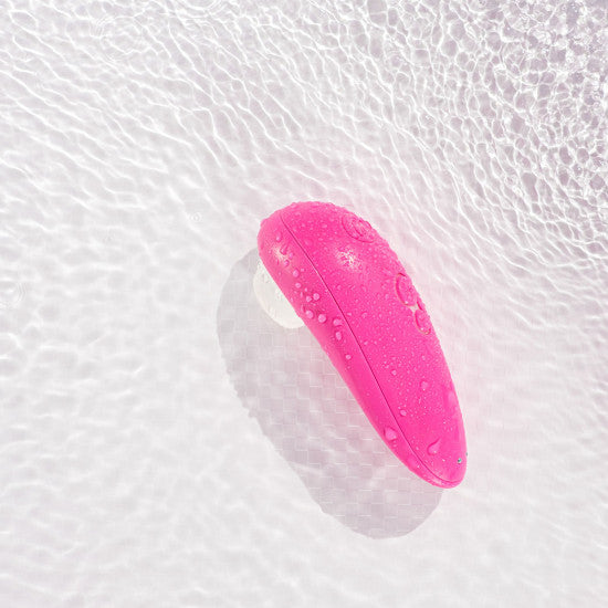 A pink, elliptical-shaped Womanizer Starlet 3 Pleasure Air Clitoral Stimulator from the Womanizer brand, with water droplets on its surface, is placed against a backdrop of rippling water. The object's sleek design and vibrant color stand out against the clear, textured water, highlighting its clitoral stimulator feature.