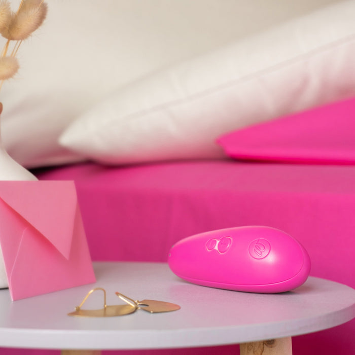 A small, pink, oval-shaped object with buttons sits on a round white table. Nearby, there's a Womanizer Starlet 3 Pleasure Air Clitoral Stimulator in pink, a pink envelope, beige dried flowers in a white vase, and a pair of gold earrings. The background features a pink bedsheet and white pillows.