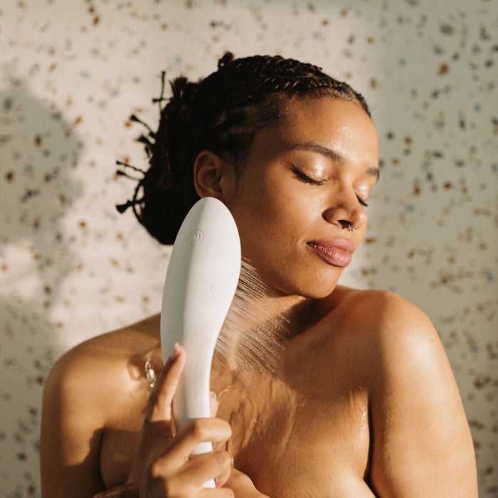 A person with braided hair stands under running water, using the Womanizer Wave Water Pulsing Shower Masturbator (White) to rinse their shoulder in a serene setting. The background features a speckled pattern, and the person has a peaceful, relaxed expression on their face as part of their self-care routine with the Womanizer brand product.