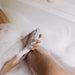 A person soaks in a bubble bath while holding the Womanizer Wave Water Pulsing Shower Masturbator (White) against their shin, likely for exfoliation or hair removal. The person's nails are neatly manicured, and the scene conveys a moment of self-care and relaxation, seamlessly fitting into their self-care routine.