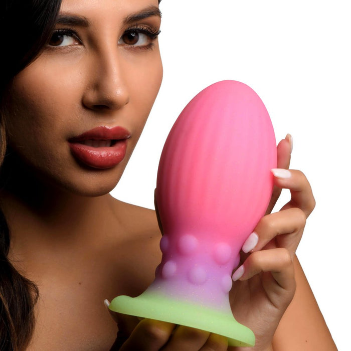 A woman with long dark hair holds the Xeno Egg Glow In The Dark Extra Large Silicone Egg from XR Brands. The cylindrical toy, made from premium silicone, features ribbed sides and a wider base adorned with raised bumps. The white background accentuates the vibrant pink and green colors of the toy prominently.