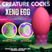 Promotional image for the "Xeno Egg Glow In The Dark Extra Large Silicone Egg" by XR Brands. This pink egg with a green base, crafted from premium silicone, features ribbed sides and a strong suction base. Set against a vibrant, otherworldly landscape, bold text highlights the product's unique attributes.