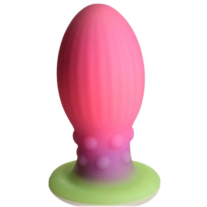 Introducing the Xeno Egg Glow In The Dark Extra Large Silicone Egg by XR Brands: a silicone butt plug with ribbed sides and a bulbous top. Featuring a gradient from pink to purple on the base, transitioning to a green flared bottom adorned with small textured bumps, this premium silicone toy glows in the dark for an added touch of excitement.