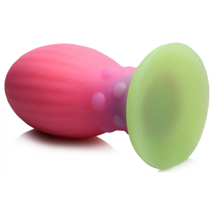 The Xeno Egg Glow In The Dark Extra Large Silicone Egg by XR Brands is displayed lying on its side. It features a pink, tapering bulbous end with slightly textured, ribbed sides and a smooth, light green flared base that glows in the dark.