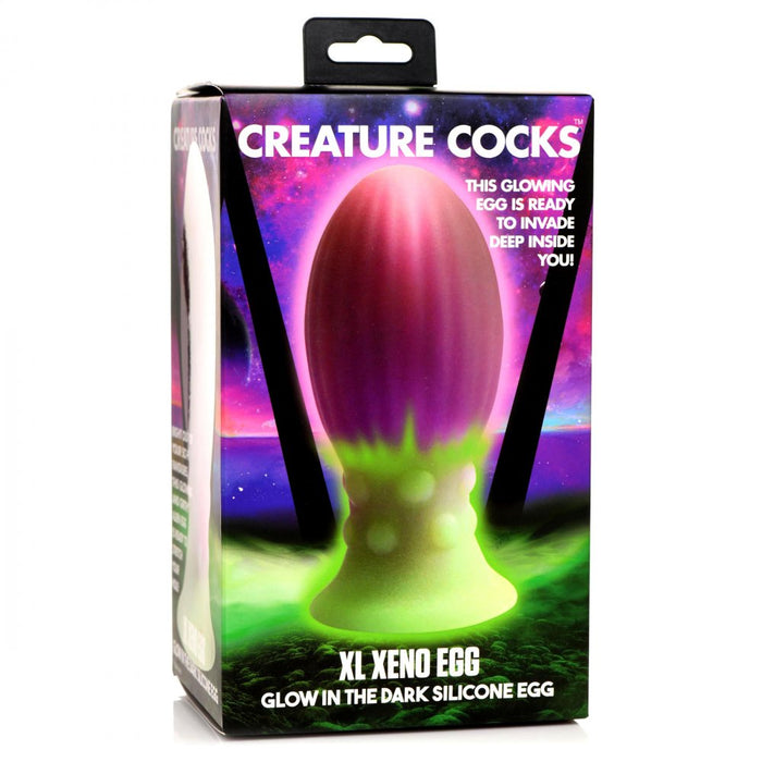 A colorful box showcases the "Xeno Egg Glow In The Dark Extra Large Silicone Egg" from XR Brands. The packaging highlights a brightly colored, glow-in-the-dark, alien-themed silicone toy with ribbed sides for added texture. The background features a cosmic, vibrant purple and green gradient with a space motif.