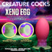 Advertisement image of a product called "Xeno Egg Glow In The Dark Silicone Egg" by XR Brands. Showcases a squishy, flexible, glow-in-the-dark egg with a beaded texture and a strong suction base. The background features a colorful, alien-like landscape. Text emphasizes its phthalate-free, super soft feel and premium silicone material.