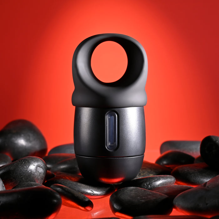 An elegant black Wet It Be Hand Held Vibrating Masturbator with Lube Dispenser by Evolved Novelties, featuring a circular handle on top, is nestled among smooth black stones. The vibrant red background creates a striking contrast with the dark tones of both the device and the stones.