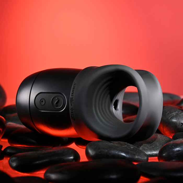 A Wet It Be Hand Held Vibrating Masturbator with Lube Dispenser from Evolved Novelties, featuring buttons on its side and a cylindrical shape with a hollow, textured inner chamber, rests on smooth black stones against a red gradient background. The device also includes a flexible silicone cradle, making it the perfect Handy Helper.