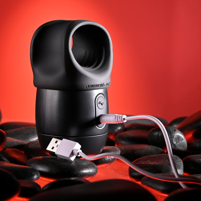 A Wet It Be Hand Held Vibrating Masturbator with Lube Dispenser by Evolved Novelties, in sleek black and cylindrical design, with an open top and a USB charging cable plugged into its side, set against a bright red background adorned with black stones arranged at the base.