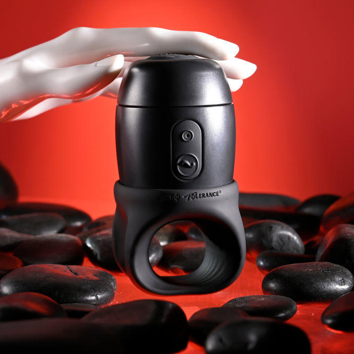 A black cylindrical Wet It Be Hand Held Vibrating Masturbator with Lube Dispenser from Evolved Novelties, featuring buttons and a ring at the bottom, sits on a surface scattered with black stones. A white mannequin hand gently touches the top of the device, all set against a vivid red background.