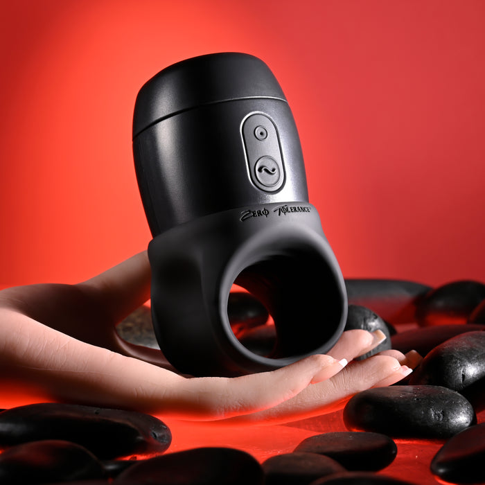 A hand holds the sleek, black Wet It Be Hand Held Vibrating Masturbator with Lube Dispenser by Evolved Novelties. The background is red with smooth black stones scattered around. This waterproof and ergonomic gadget features adjustable vibrating speeds and a convenient loop at the bottom.