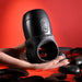 A hand holds the sleek, black Wet It Be Hand Held Vibrating Masturbator with Lube Dispenser by Evolved Novelties. The background is red with smooth black stones scattered around. This waterproof and ergonomic gadget features adjustable vibrating speeds and a convenient loop at the bottom.