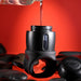 A Wet It Be Hand Held Vibrating Masturbator with Lube Dispenser from Evolved Novelties, black and cylindrical with a ring handle, is being filled with liquid from a small bottle against a red background. The device is placed among several smooth black stones, with one cap labeled "PUMP" resting beside it.