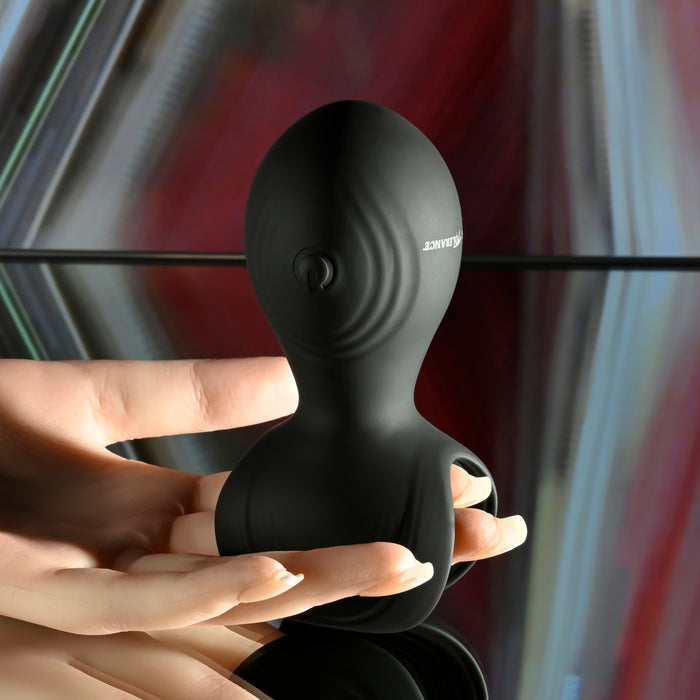 A hand holds the Handy Helper Petite Hand Held Vibrating Masturbator by Evolved Novelties, a black, uniquely shaped object with a smooth surface against a blurred background featuring metallic and colorful hues. The masturbator's design, reminiscent of a flexible silicone cradle, includes curved contours and a circular button on one side.