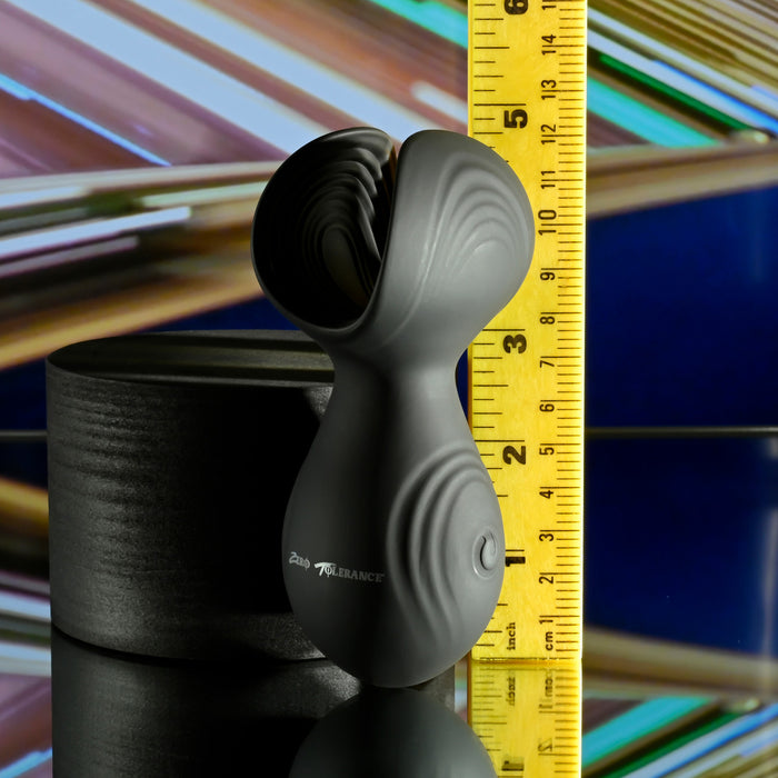 The Handy Helper Petite Hand Held Vibrating Masturbator by Evolved Novelties, a sleek black abstract-shaped object, stands upright next to a yellow measuring tape, showing a height of about 4 inches. The background features colorful, blurred lines, and part of a cylindrical black container can be seen. Its hand-held size design includes a flexible silicone cradle for added convenience.