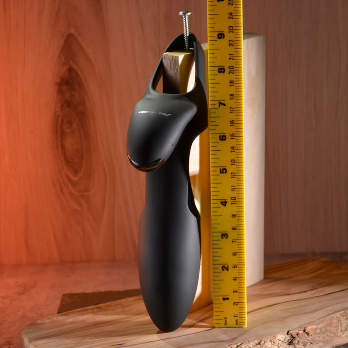 A black Nautilus Rechargable Vibrating Penis Sleeve, resembling a sleek design with mounted screw and remote control, rests on a wooden surface beside a yellow ruler. Warm lighting enhances the ambiance against the wooden background.