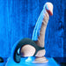 A realistic dildo with a harness, illuminated by blue lighting, stands on a smooth surface against a deep blue wooden background. The scene, which has a clinical yet atmospheric feel, also features the Zero Tolerance Vibrating Black Ball Cradle by Evolved Novelties that seamlessly complements the setting.