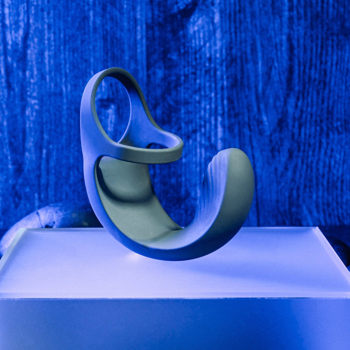 A modern, abstract sculpture with smooth, curved lines and an open circular design, standing on a sleek, illuminated white platform against a blue textured background. The artwork features a gradient of green shades and appears to float effortlessly like the Zero Tolerance Vibrating Black Ball Cradle by Evolved Novelties.