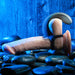 The Zero Tolerance Vibrating Black Ball Cradle by Evolved Novelties, featuring a realistic silicone strap-on and a black harness, is elegantly positioned on a smooth stone surface. Bathed in an ambient blue glow, the scene exudes a moody, intimate atmosphere. The included remote control vibrator enhances the experience, offering unparalleled pleasure and convenience.