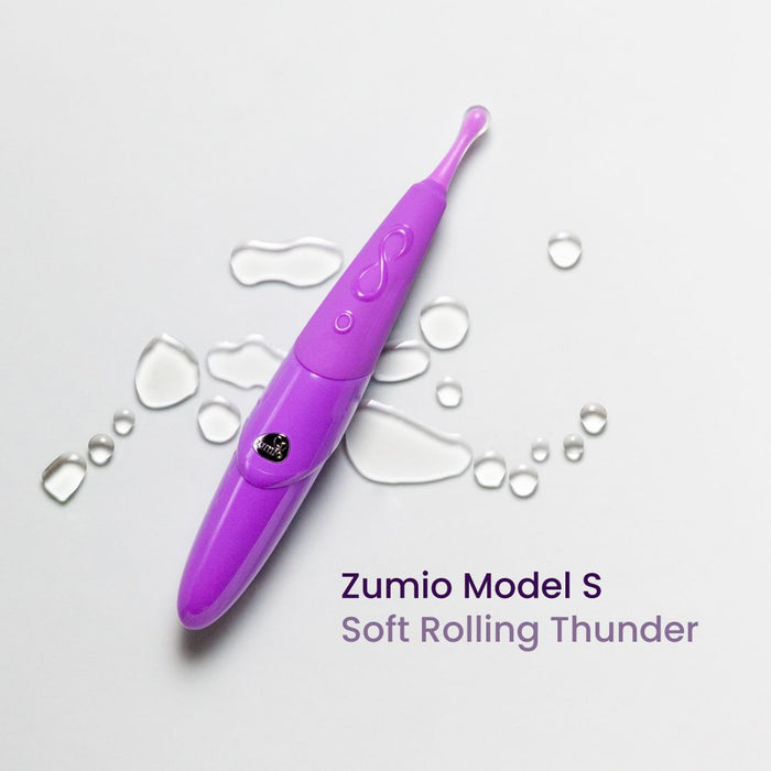 A purple Zumio S rechargeable clitoral stimulator with a curved, oval SpiroTIP™, displayed on a white background. Water droplets surround the device. Text reads: "Zumio S Soft Rolling Thunder, for precise pinpoint stimulation.