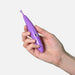 A hand holding the Zumio S - Rechargeable Clitoral Stimulator, a small purple device featuring a rounded SpiroTIP and a silver button on its side. The Zumio, crafted for unique stimulation, appears to be made of plastic, with the hand positioned against a plain white background.