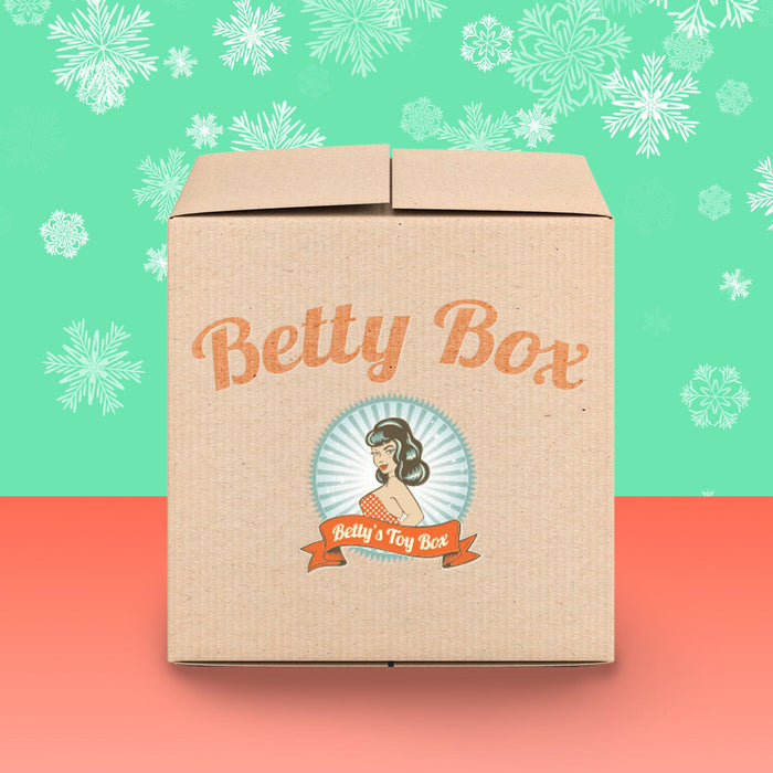 A cardboard box labeled "Betty's Kinky Kitties Mystery Box - Holiday 2024 Edition" by Betty's Toy Box, adorned with vintage-style graphics, piques curiosity as it rests on a two-toned background of green with snowflake patterns and a coral base, hinting at the enticing treats within the mystery package.