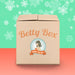 A cardboard box labeled "Betty's Kinky Kitties Mystery Box - Holiday 2024 Edition" by Betty's Toy Box, adorned with vintage-style graphics, piques curiosity as it rests on a two-toned background of green with snowflake patterns and a coral base, hinting at the enticing treats within the mystery package.