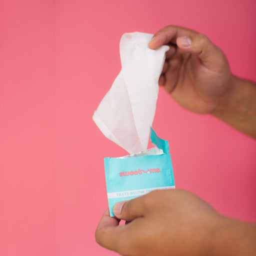 Two hands hold an open package of Sweetums Flavored Intimate Wipes, featuring a turquoise blue and white design against a pink background. Made from plant fibers, one hand smoothly pulls a single white wipe from the pH-balanced pack.