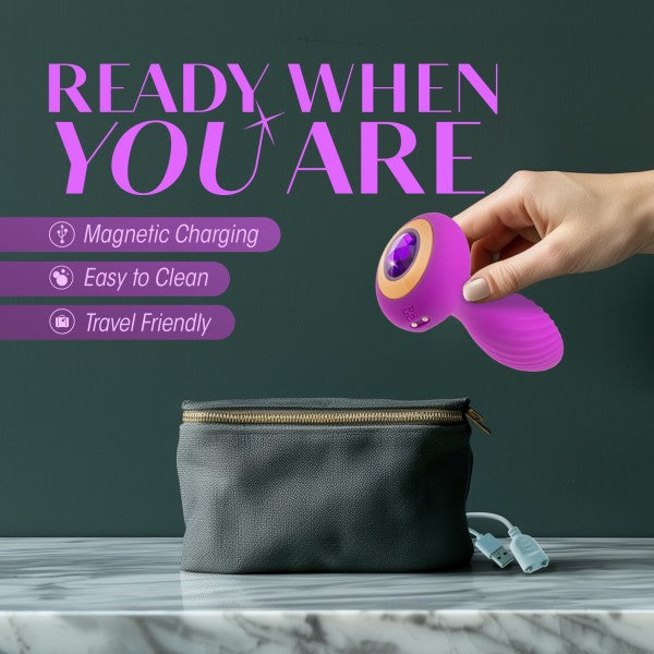 A hand holding a vibrant amethyst Charm Vibrating Textured Silicone Butt Plug by Blush, next to a black toiletry bag on a marble surface. Text above reads, "READY WHEN YOU ARE" and lists features: Magnetic Charging, Easy to Clean, Travel Friendly with powerful vibrations.