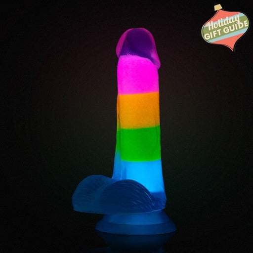 The Blush Rainbow Rave Glow in the Dark 6.5 Inch Silicone Dildo stands upright against a dark background, highlighting its playful and vibrant multicolored design.