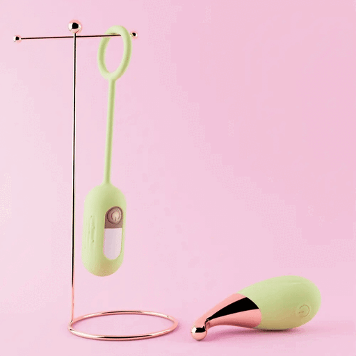 A pair of pastel green and rose gold-colored electronic personal items (Blush Blooming Bliss First Time Bullet & Egg Vibrators with Charging Case & Remote) on a pink background. One item, which includes a wireless remote control, hangs on a minimalist metal stand with a circular base, while the other sits next to it, showing its rounded tip and button.