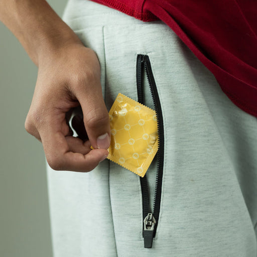 A person in light gray pants holds a Royal Condom Tailored Fit Vegan Condom from the yellow 20-pack near the zippered pocket, partially visible with the edge of a red shirt showing.