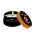 The Pumpkin Spice Erotic Massage Candle - Your Trick, My Treat by Kama Sutra, made with coconut oil, sits in a sleek black container. Its orange-rimmed lid, adorned with a pumpkin design and the playful text "Your trick... my treat!" lies beside it. The candle's flame glows softly, casting a warm light and releasing the inviting scent of pumpkin spice into the air.